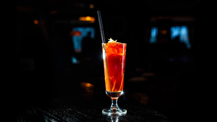 A red cocktail in a tall glass stands on the counter of a dark bar.