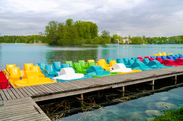 A wooden pier near lakeshore with the row of colored plastic catamarans with lake water and forest...