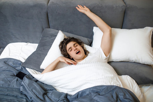 Image of a handsome young man in the morning yawning and stretching in modern bed