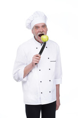 Old man, professional chef in white uniform holding metal knife with green apple while staying on white background