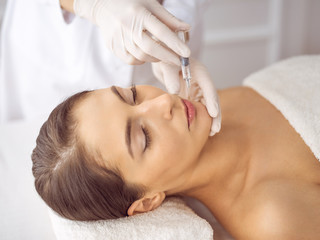 Fototapeta na wymiar Beautician doing beauty procedure with syringe to face of young brunette woman. Cosmetic medicine and surgery, beauty injections
