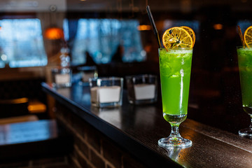 A green cocktail in a tall glass stands on the counter of a dark bar.