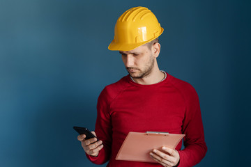 Portrait of young engineer or building contractor wearing yellow protective helmet red sweater standing in front of blue wall background holding smart phone and clipboard reading messages or report