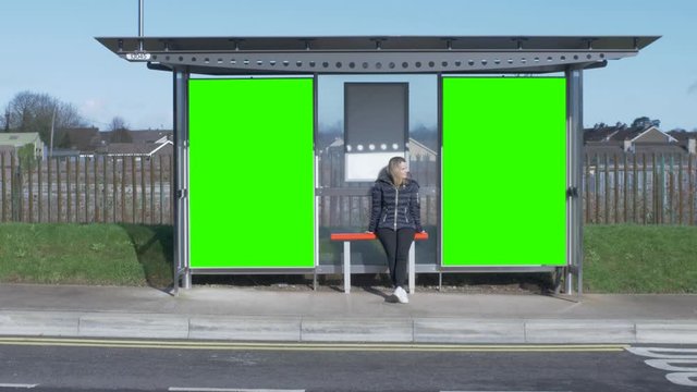 Attractive young woman waits for the bus at a bus stop sitting. Green Screen Posters - Track the video and add your own advertisement.