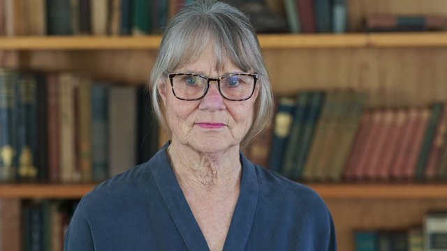 Medium close up portrait of senior Caucasian woman in front of book case at home or library. Older white lady looking at camera in home office. Slow motion 4k