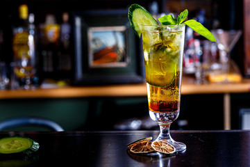 A green cocktail or lemonade with cucumber in a tall glass stands on the counter of a dark bar.