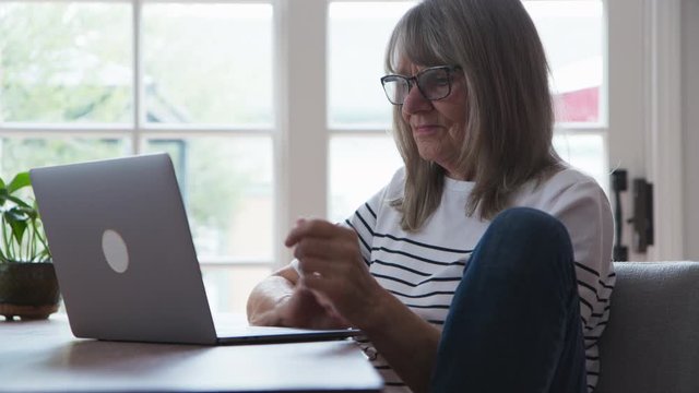 Senior Caucasian woman sitting casually with leg up working on laptop computer at dining room table. Older white lady working at home on pc. Slow motion 4k