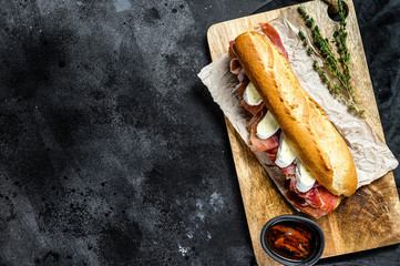 baguette sandwich with jamon ham serrano, paleta iberica, Camembert cheese on the cutting Board.  Black background, top view, space for text - obrazy, fototapety, plakaty