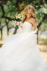 Fototapeta na wymiar Cute young bride with long hairs holding her wedding bouquet includes white roses and other flowers. Beautiful white marriage dress. Pretty girl on greet trees background