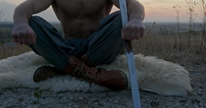 A lonely and powerful warrior meditates with a sword on a high mountain against a sunset background