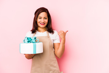 Middle age latin woman holding a cake isolated on a pink background smiling and raising thumb up