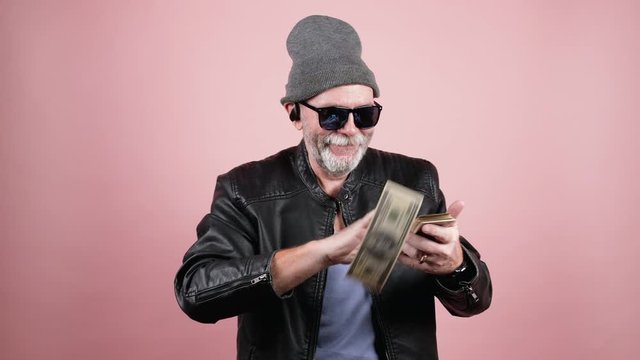 Portrait of a modern senior hipster man in leather jacket, grey hat, sunglasses and earpods throwing dollar banknotes from the stack and laughing.  Finance, rich people and modern lifestyle concept. 