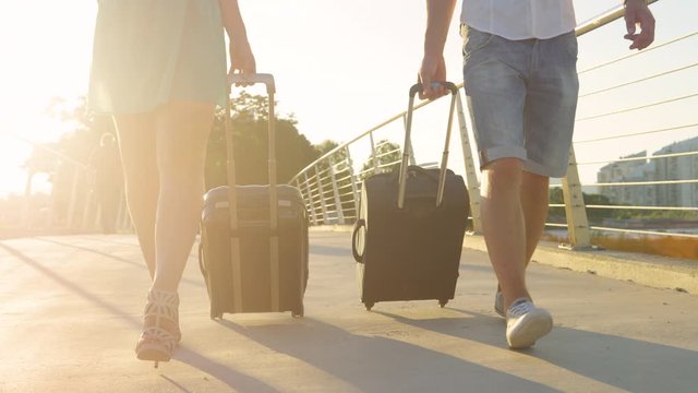 SLOW MOTION, LOW ANGLE, CLOSE UP, LENS FLARE: Cinematic shot of a tourist couple dragging their suitcases to the airport. Unrecognizable woman in high heels and boyfriend pull luggage to the airport.