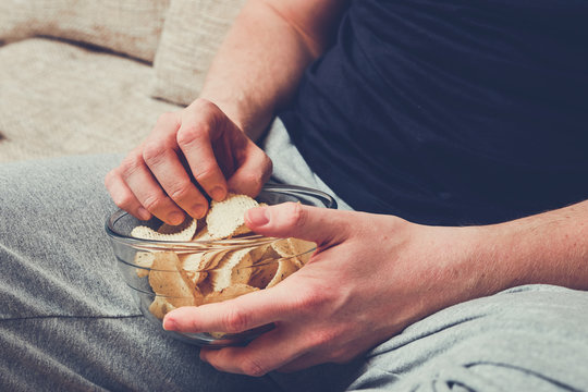 A Man Sits On A Sofa And Eats Chips From A Glass Dish.