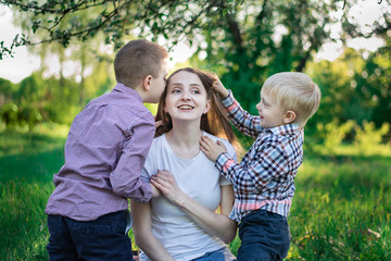 Fototapeta na wymiar Young pretty mother with two sons sit in the park. Children kiss and hug mom