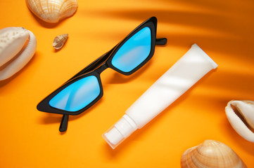 Fashionable glasses and a tube of moisturizer or sunscreen on a yellow background with sea shells and shade from a palm leaf. The concept of self-care , sun protection and safe rest.