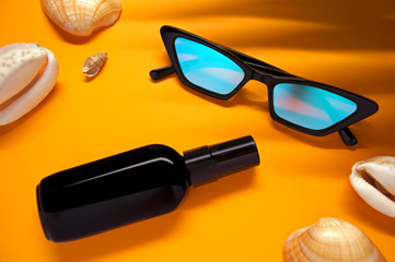 Top view of fashionable glasses and a bottle of moisturizer or sunscreen on a yellow background with seashells and shade from a palm leaf.