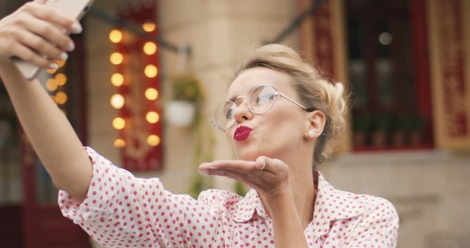 Pretty Caucasian girl smiling and making selfie photo on phone camera in city. Close up portrait of beautiful 80s retro style lady in glasses posing on street. Cheerful red lips blonde female model
