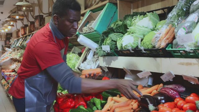 Confident African American seller in apron arranging fresh vegetables and fruits on greengrocery counter
