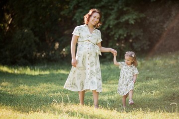 Naklejka na ściany i meble A beautiful young pregnant mom and her little daughter are picking flowers in a field. Motherhood. A family.