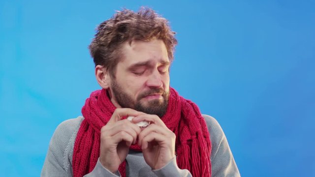 Unhealthy allergic young bearded man sneezing and cleaning nose snot with paper napkin. Sick isolated man on blue background. Medicine, healthcare concept.