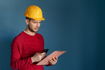 Portrait of young engineer or building contractor wearing yellow protective helmet red sweater standing in front of blue wall background holding smart phone and clipboard reading messages or report