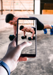 hands picking up a smartphone taking a picture of a young man doing exercise in exterior. Space for text. Selective focus