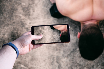 hands picking up a smartphone taking a picture of a young man doing exercise in exterior. Space for text. Selective focus