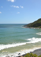 Waves are breaking onto a rocky beach, which leads to a tree-covered headland. Two surfers are barely visible in the water. The sky is blue with faint clouds.