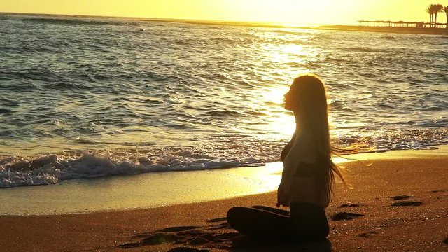 Yoga by sea and woman with hands up meditation in sun salut on lotus position at seaside with coming sea waves in sun light sunrise.