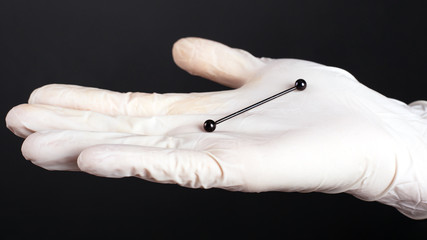 hand in a white glove holds a cartilage piercing ear jewelry black color on a dark background...