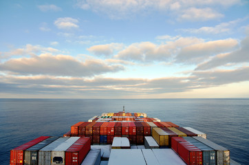 View on the containers loaded on deck of the large cargo ship. She is sailing through calm, blue ocean.  - obrazy, fototapety, plakaty