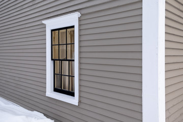 The exterior wall of a grey colored building made of narrow wooden clapboard with a large drift of snow. There's a double hung closed window in the wall with white trim and black glass dividers.