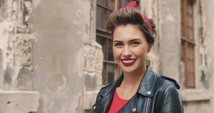 Charming happy trendy girl standing in the street. Young pretty vintage woman in leather jacket with smile on face. American retro style female model with red lips posing outdoors. Fashion concept