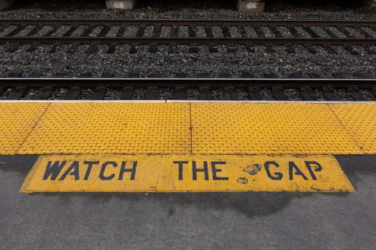 sign at the railroad station at the edge of the platform looking toward the tracks warning you to watch the gap