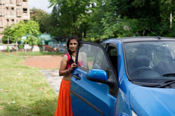 Young and attractive Indian Bengali brunette woman trying to get out of a blue car by opening door wearing Indian traditional ethnic cloths pink and orange skirt blouse. Indian lifestyle and fashion