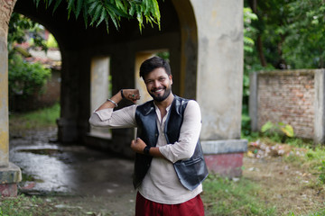 portrait of young and handsome Indian Bengali brunette beard man posing in green nature in front of vintage house wearing Indian ethnic vibrant jacket, shirt and red pant. Indian lifestyle and fashion - Powered by Adobe