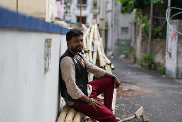 Obraz premium Portrait of an young and handsome Indian Bengali brunette beard man sitting on a pile of bamboos on roadside wearing black jacket and white shirt. Indian lifestyle and fashion