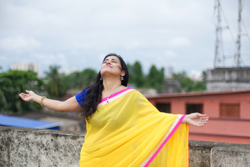 Fototapeta premium Young and beautiful Indian Bengali brunette woman in Indian traditional yellow sari and blue blouse is standing while spreading her arms on rooftop under blue sky with clouds. Indian lifestyle