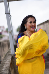 Young and beautiful Indian Bengali brunette woman in Indian traditional dress yellow sari and blue blouse standing on rooftop under blue sky with clouds with her sari blown in air. Indian lifestyle