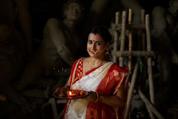 An young and beautiful Indian Bengali brunette woman in red white traditional ethnic sari holding a puja thali for worshiping goddess Durga clay idol . Indian culture, religion and fashion