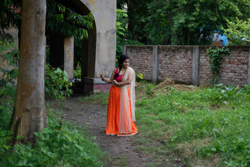 Young and beautiful Indian Bengali brunette woman standing thoughtfully in green nature in front of a vintage house wearing Indian traditional ethnic vibrant skirt blouse. Indian lifestyle and fashion
