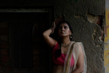 Fototapeta premium Young and attractive Indian Bengali brunette woman standing in front of a vintage house window wearing Indian traditional ethnic cloths pink and orange skirt blouse. Indian lifestyle and fashion