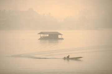Small boat approached raft house floating in middle of a lake. before sunrise moment in fog rural river. Thailand outback vacation destination. romantic peaceful scene of slow life living in nature.