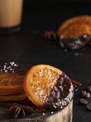Christmas sweets, candied fruits in chocolate. Caramelized orange slices on a wooden stand, black stone background. Nearby is a cup with cappuccino, spices and chocolate. Homemade dessert. Close-up.