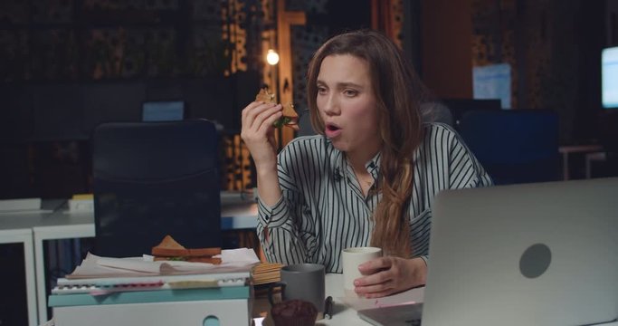 Sad businesswoman having late meal while sitting at desk with open laptop in front of her. Tired girl employee eating sandwich and drinking while working long hours in empty office.