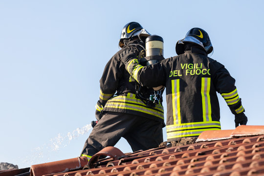 Close up of two Italian fire fighters standing on a roof and handling a hose to extinguish a smoldering tile