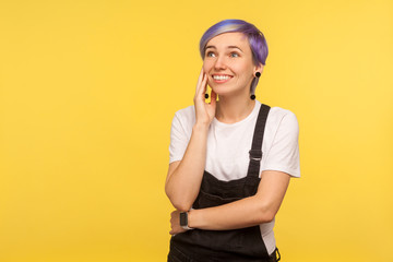 Portrait of happy beautiful hipster girl with violet short hair in denim overalls holding palm near face and dreaming pleasant fantasies, sweet memories. isolated on yellow background, studio shot