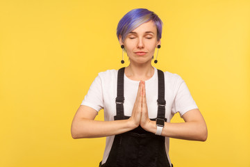 Yoga meditation, harmony. Portrait of peaceful hipster woman with violet short hair in denim overalls holding palms together in namaste or prayer gesture. isolated on yellow background, studio shot