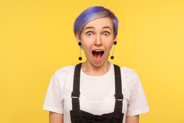 Wow, unbelievable! Portrait of amazed hipster woman with violet short hair in denim overalls looking surprised with open mouth, expressing excitement, shock. isolated on yellow background, studio shot